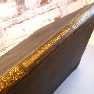 Op de afbeelding: Een antiek boek met een donkere kaft en gouden letters. De titel "Histoire de Jules César Atlas" is zichtbaar op de rug, samen met decoratieve gouden filigraan. Het boek lijkt oud, met een getextureerd oppervlak.