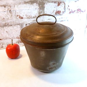Könnte beinhalten: Ein Vintage-Metallbehälter mit Deckel und Griff. Der Behälter hat eine zylindrische Form mit leicht ausgestelltem Boden und einem angelaufenen, gealterten Aussehen. Links vom Behälter liegt ein roter Apfel.