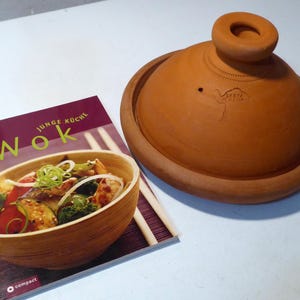 Könnte beinhalten: Ein Terrakotta-Tajine-Topf mit konischem Deckel und kleinem Griff. Der Topf steht neben einem Kochbuch mit dem Titel "Wok" mit einem Foto eines Pfannengerichts. Der Einband ist bordeauxfarben mit den Worten "Junge Küche" über "Wok".