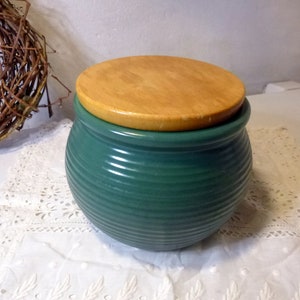 Puede incluir: Un tarro de cerámica verde con tapa de madera. El tarro tiene un diseño acanalado y está sobre un mantel de encaje blanco.