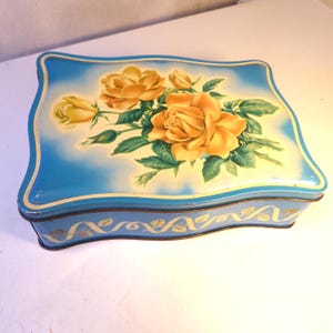 Può includere: Una scatola di metallo blu vintage con parte superiore e inferiore curve. Il coperchio presenta un motivo floreale di rose gialle e foglie verdi su uno sfondo azzurro. La scatola ha finiture dorate e un motivo dorato decorativo attorno alla base.