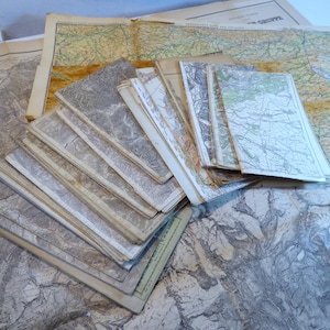 Op de afbeelding: Een verzameling vintage kaarten, sommige opgevouwen en opgestapeld, andere uitgespreid. De kaarten bevatten gedetailleerde geografische informatie met verschillende kleuren en tinten. Het kleurenpalet is over het algemeen gedempt, met tinten beige, grijs en groen.