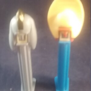 May include: Two PEZ dispensers. One is white with a gold accent on top. The other is blue with a yellow, round head. Both have a rectangular base and are standing upright.