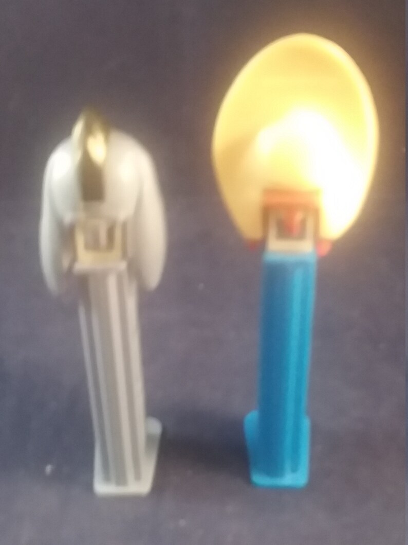 May include: Two PEZ dispensers. One is white with a gold accent on top. The other is blue with a yellow, round head. Both have a rectangular base and are standing upright.