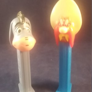 May include: Two vintage PEZ dispensers. One is a gray donkey with a sad expression, and the other is a cartoon character with a yellow head, red mustache, and blue base. Collectible candy dispensers.