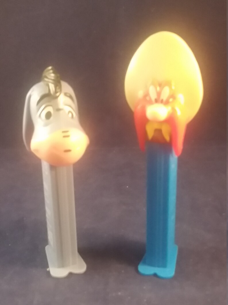 May include: Two vintage PEZ dispensers. One is a gray donkey with a sad expression, and the other is a cartoon character with a yellow head, red mustache, and blue base. Collectible candy dispensers.