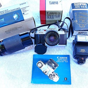 Puede incluir: Una cámara de película Canon AV-1 plateada con un objetivo Canon FD 50 mm f/1,8 montado. La cámara está sobre una superficie blanca con un objetivo negro, un flash y un manual de instrucciones de la cámara. El flash es un Sunpak auto 144.