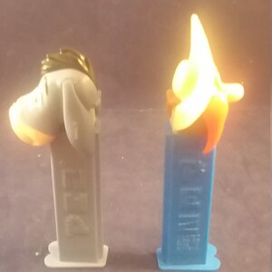 May include: Two PEZ dispensers. One is gray with a donkey head, the other is blue with a star-shaped head. Both have the PEZ logo on the dispenser body. The donkey has a light orange muzzle and the star has orange and red accents.
