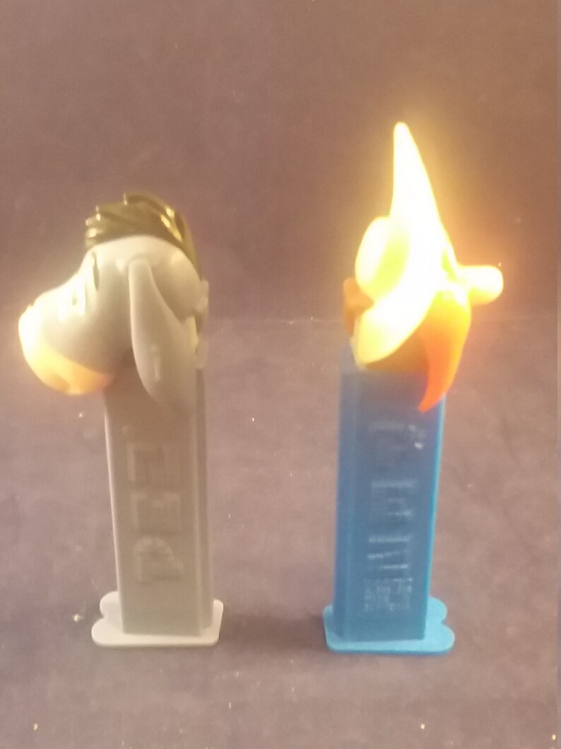 May include: Two PEZ dispensers. One is gray with a donkey head, the other is blue with a star-shaped head. Both have the PEZ logo on the dispenser body. The donkey has a light orange muzzle and the star has orange and red accents.