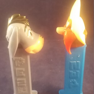 May include: Two PEZ dispensers featuring cartoon characters. One is gray with a donkey head, the other is blue with a character with a large white hat and orange face. The dispensers are side-by-side.