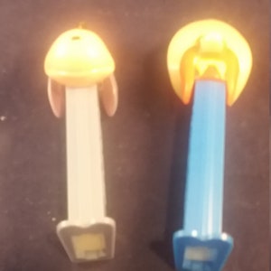 May include: Two PEZ candy dispensers. One has a yellow head with pink ears and a light gray body. The other has a yellow head and a blue body. Both have a rectangular base.