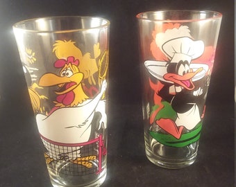 May include: Two vintage drinking glasses featuring cartoon characters. The first glass depicts a yellow rooster playing badminton. The second glass shows a black duck wearing a chef's hat and holding a spatula.