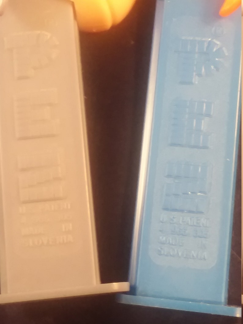 May include: Close-up of two PEZ dispensers. One is light beige, the other is light blue. Both have the PEZ logo and text that reads "U.S. Patent" and "Made in Slovenia".