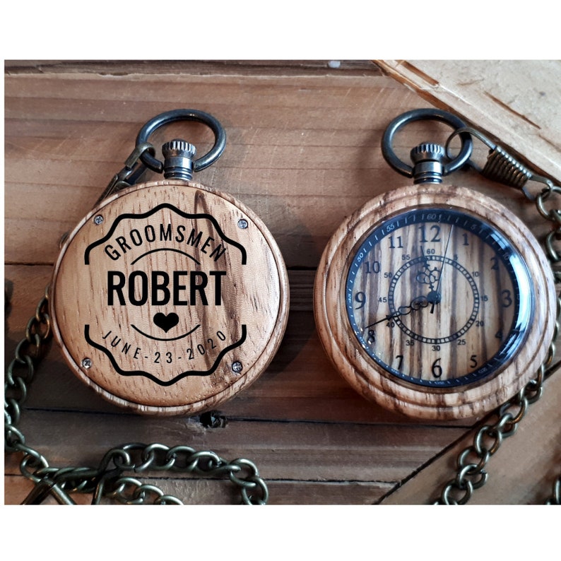 May include: Two wooden pocket watches with a chain. One watch has a black and white engraved design that says "Groomsmen Robert June 23-2020". The other watch has a traditional clock face.