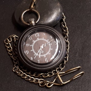May include: A black pocket watch with a gold chain and a wooden face. The watch face has white Roman numeral hour markers and black hands. The watch is on a black surface.