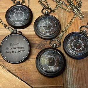 May include: Five antique-style pocket watches with chains. Four of the watches have Roman numeral faces and the fifth watch has an engraved message that reads "Shawn Groomsman July 23, 2022".