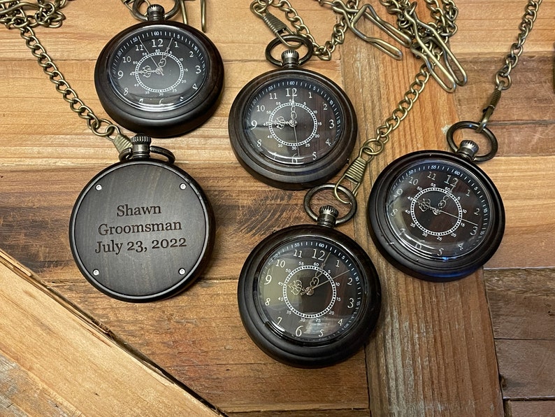 May include: Five antique-style pocket watches with chains. Four of the watches have Roman numeral faces and the fifth watch has an engraved message that reads "Shawn Groomsman July 23, 2022".