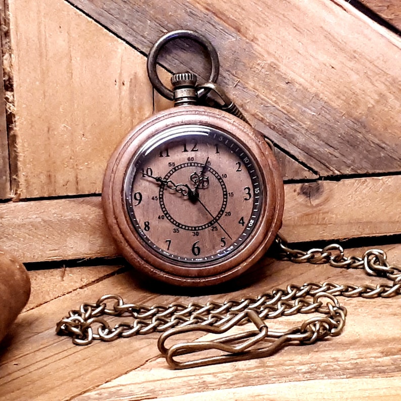May include: A brown wooden pocket watch with a brass chain. The watch face has Roman numerals and a black hand. The watch is on a wooden surface.
