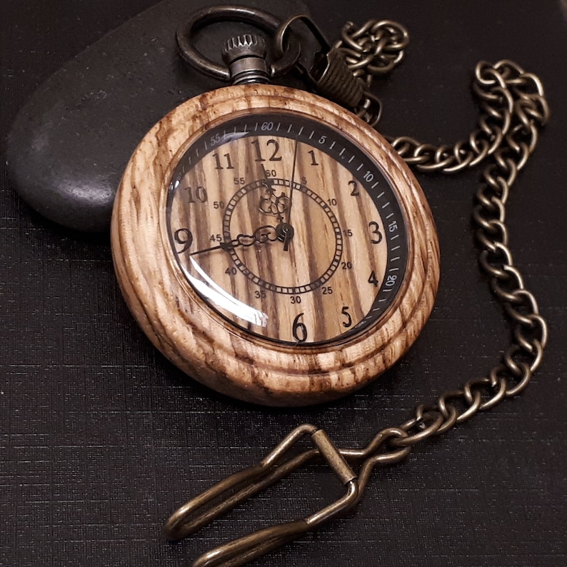 May include: A wooden pocket watch with a brown wood grain finish and a clear face with black Roman numeral markings. The watch has a brass chain attached.