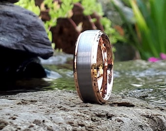 May include: A silver and rose gold wedding band with a brushed finish. The band is shown on a rock near a stream.