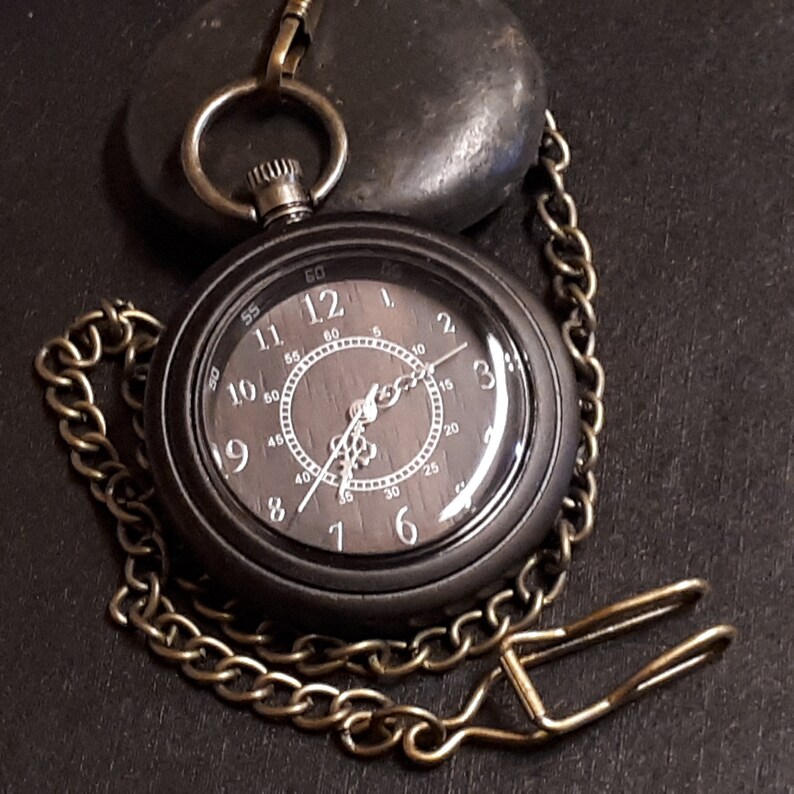 May include: A black pocket watch with a gold chain and a wooden face. The watch face has white Roman numeral hour markers and black hands. The watch is on a black surface.