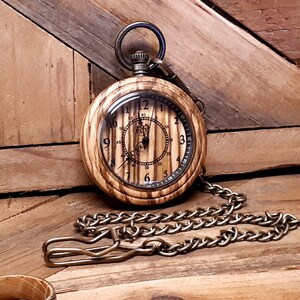 May include: A wooden pocket watch with a brass chain. The watch face has a wood grain design and Roman numeral markings. The watch is on a wooden surface.