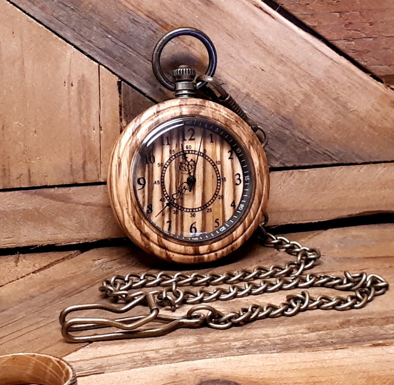 May include: A wooden pocket watch with a brass chain. The watch face has a wood grain design and Roman numeral markings. The watch is on a wooden surface.