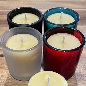 May include: Four candles in glass jars with different colored glass. The candles are white and have wicks in the center. The jars are clear, black, teal, and red.