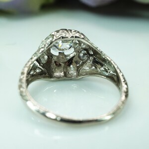 May include: A vintage-style silver ring with a large, clear, round-cut center stone and smaller accent stones. The ring features intricate filigree detailing around the setting and band, showcasing a classic, elegant design. The ring is set on a white surface.