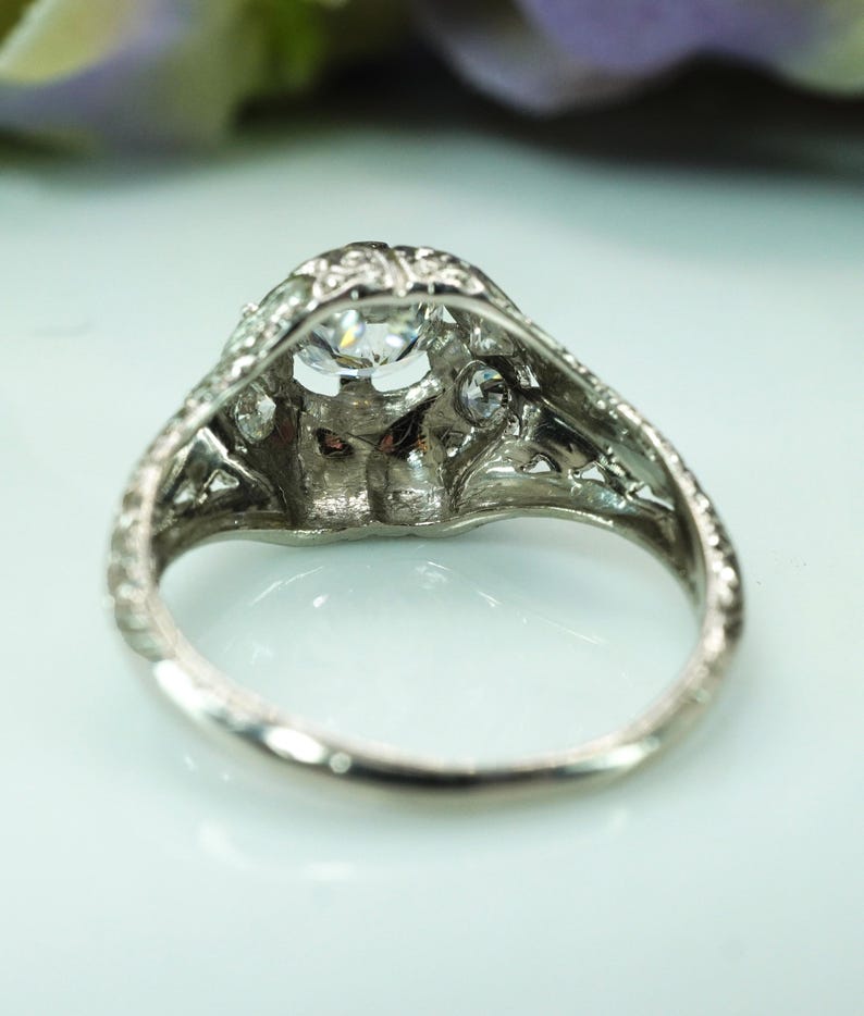 May include: A vintage-style silver ring with a large, clear, round-cut center stone and smaller accent stones. The ring features intricate filigree detailing around the setting and band, showcasing a classic, elegant design. The ring is set on a white surface.