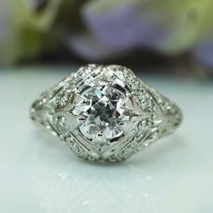 May include: A close-up of a vintage-style ring with a large, clear, round-cut diamond as the centerpiece. The ring is made of white metal, possibly platinum or white gold, and features intricate filigree and small accent diamonds around the main stone.