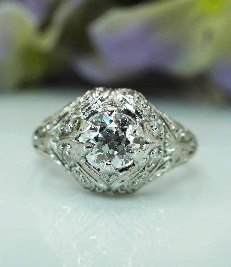 May include: A close-up of a vintage-style ring with a large, clear, round-cut diamond as the centerpiece. The ring is made of white metal, possibly platinum or white gold, and features intricate filigree and small accent diamonds around the main stone.
