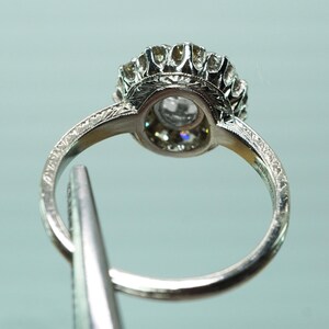 May include: Close-up of a silver ring with a large, clear gemstone at the center, surrounded by smaller stones. The ring has an ornate, detailed design with etched patterns on the band. The ring is held by a silver tool.
