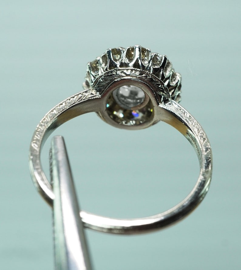 May include: Close-up of a silver ring with a large, clear gemstone at the center, surrounded by smaller stones. The ring has an ornate, detailed design with etched patterns on the band. The ring is held by a silver tool.