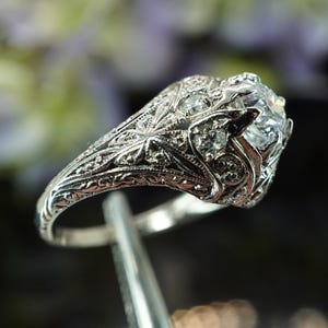 May include: A detailed, silver-toned ring with a large, clear gemstone at the center. The ring features intricate filigree and small accent stones. The design is ornate and vintage-inspired, with a focus on craftsmanship and detail.