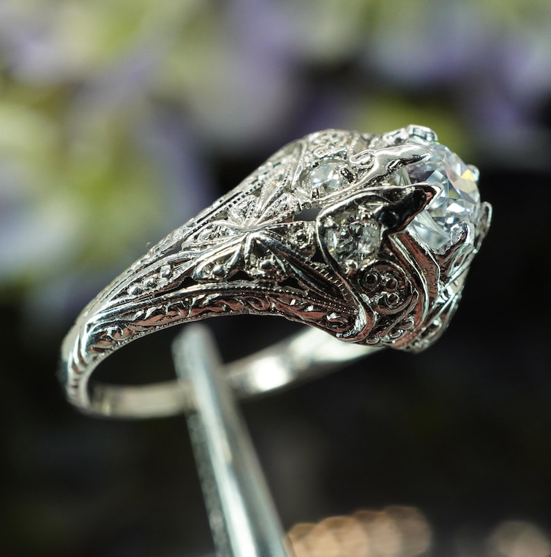 May include: A detailed, silver-toned ring with a large, clear gemstone at the center. The ring features intricate filigree and small accent stones. The design is ornate and vintage-inspired, with a focus on craftsmanship and detail.