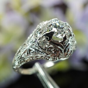 May include: A detailed, silver-toned ring with a large, clear gemstone at its center. The ring features intricate filigree and floral designs, showcasing a vintage or antique style. The gemstone is held in place by delicate prongs.