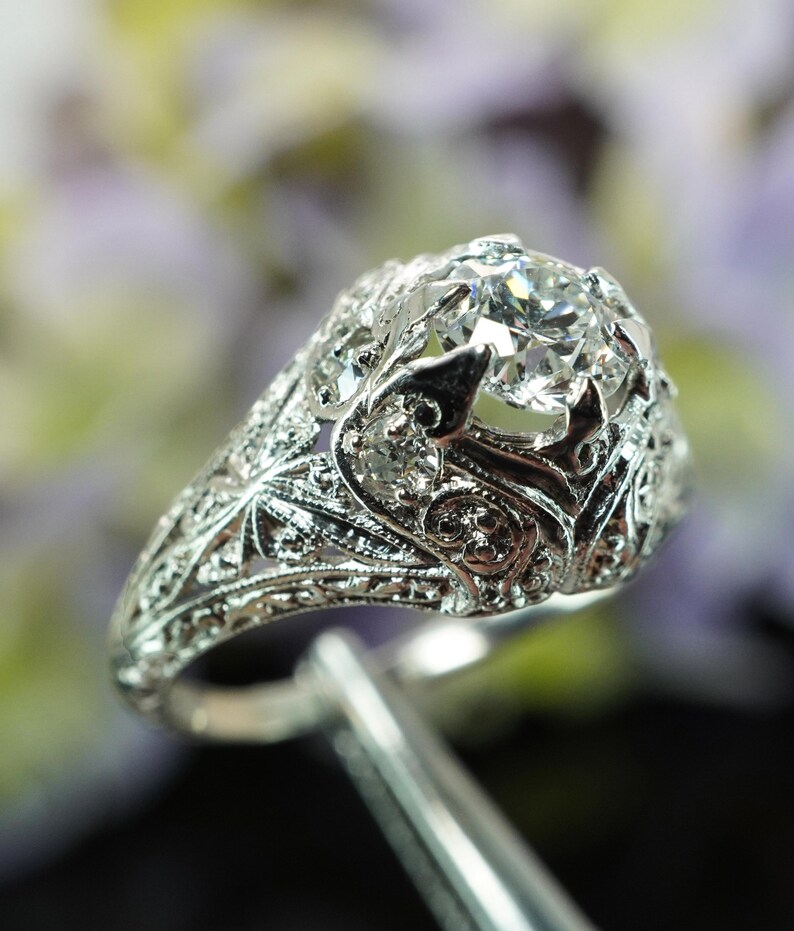 May include: A detailed, silver-toned ring with a large, clear gemstone at its center. The ring features intricate filigree and floral designs, showcasing a vintage or antique style. The gemstone is held in place by delicate prongs.