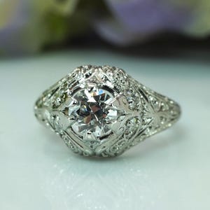 May include: A close-up of a vintage-style ring with a large, clear, round-cut diamond as the centerpiece. The ring is made of white metal, possibly platinum or white gold, and features intricate filigree detailing around the diamond and along the band.