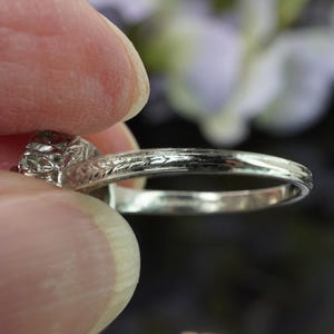 May include: Close-up of a silver ring with a detailed floral design on the band and a faceted gemstone. The ring is held between two fingers, showcasing the intricate details of the jewelry.