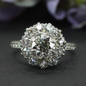 May include: A silver ring with a large, round, clear diamond center stone, surrounded by smaller diamonds in a floral halo design. The band has a textured pattern. The ring is set against a dark background.