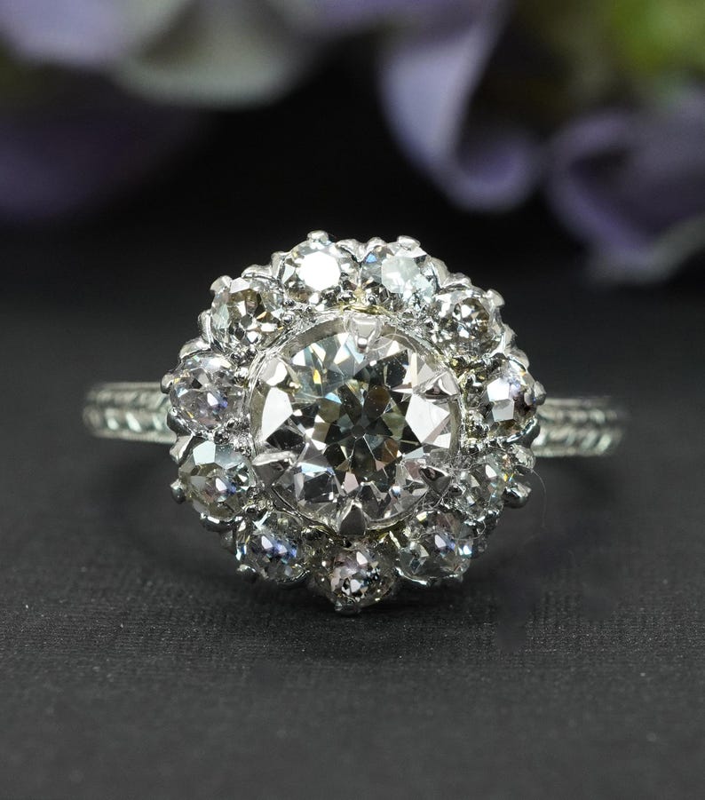 May include: A silver ring with a large, round, clear diamond center stone, surrounded by smaller diamonds in a floral halo design. The band has a textured pattern. The ring is set against a dark background.