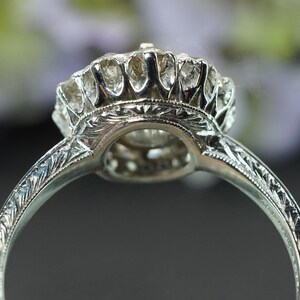 May include: Close-up of a silver ring with an intricate, engraved band. The ring features a large, round, clear gemstone surrounded by smaller stones. The setting has a detailed, floral-like design.