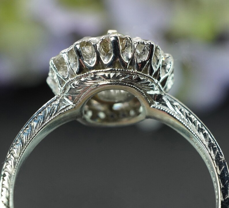 May include: Close-up of a silver ring with an intricate, engraved band. The ring features a large, round, clear gemstone surrounded by smaller stones. The setting has a detailed, floral-like design.