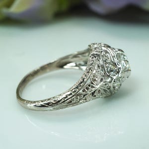 May include: Close-up of an antique silver ring with a detailed filigree design. The ring features a central cluster of small, clear gemstones, likely diamonds, set within an ornate, floral-inspired setting. The band is also intricately carved.