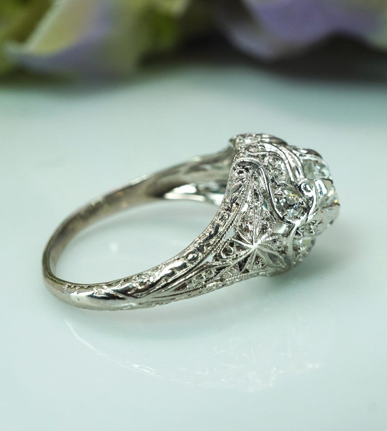 May include: Close-up of an antique silver ring with a detailed filigree design. The ring features a central cluster of small, clear gemstones, likely diamonds, set within an ornate, floral-inspired setting. The band is also intricately carved.