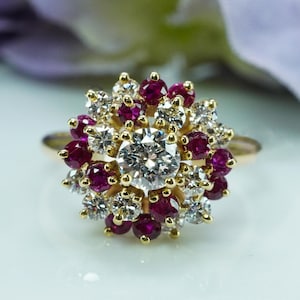 Könnte beinhalten: Ein goldener Ring mit einem zentralen, klaren Diamanten, umgeben von kleineren weißen Diamanten und tiefroten Rubinen. Die Edelsteine sind in einem floralen Muster angeordnet. Der Ring ist vor einem weichen, verschwommenen Hintergrund platziert.