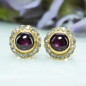May include: A pair of gold-toned stud earrings. Each earring features a deep purple, round gemstone surrounded by a halo of small, clear gemstones. The earrings are set against a soft, blurred background.