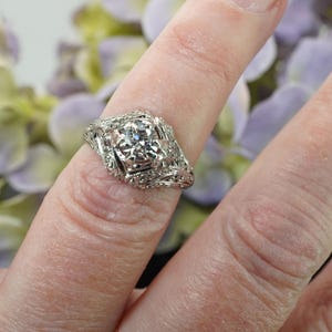 May include: A detailed, antique-style silver ring with a central, clear gemstone. The ring features intricate filigree and floral designs around the main stone and along the band. The ring is shown on a finger, with a blurred floral background.