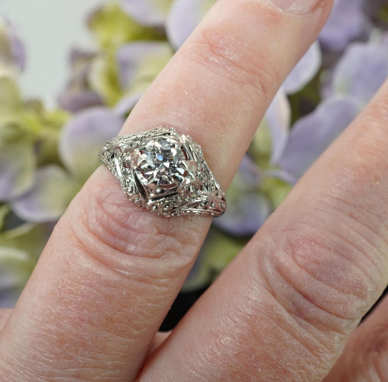 May include: A detailed, antique-style silver ring with a central, clear gemstone. The ring features intricate filigree and floral designs around the main stone and along the band. The ring is shown on a finger, with a blurred floral background.